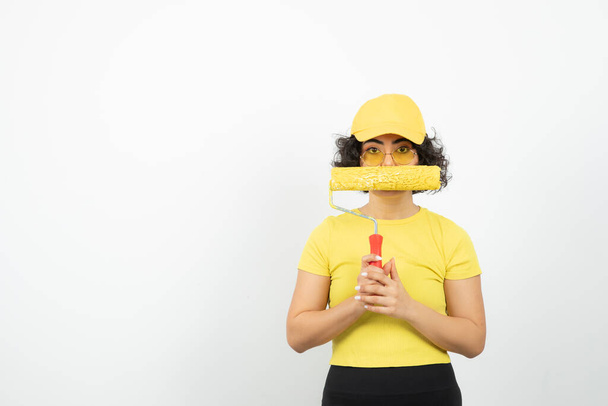 Portrait of a painter woman in cap holding paint roller over a white wall . High quality photo - Photo, Image