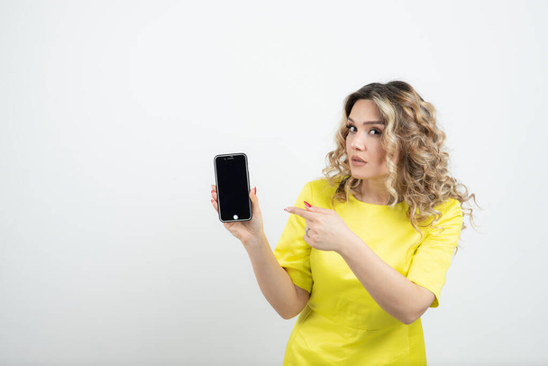 Foto di giovane donna con i capelli ondulati in abito giallo che indica il suo smartphone. Foto di alta qualità - Foto, immagini