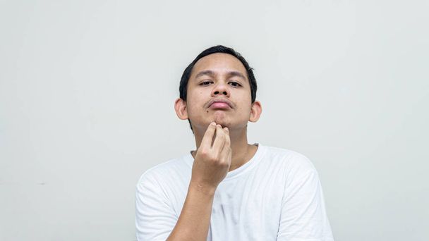A portrait of young Asian Malay man holding chin checking his face for skin care and beauty concepts, studio shot isolated white background with copy space. - Fotografie, Obrázek