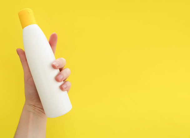 Female hand holding white cosmetic bottle on yellow background. Mock up, copy space. Summer skincare concept - Zdjęcie, obraz