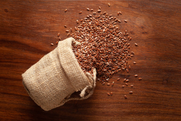 Close-up of Organic Brown flaxseeds (Linum usitatissimum) or linseed spilled out from a laying jute bag over wooden brown background. - Foto, afbeelding
