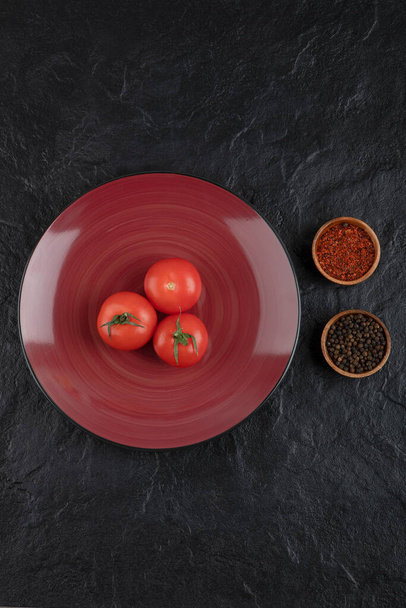 Um prato vermelho de três tomates vermelhos frescos com pimenta e pimenta em pó sobre um fundo preto. Foto de alta qualidade - Foto, Imagem