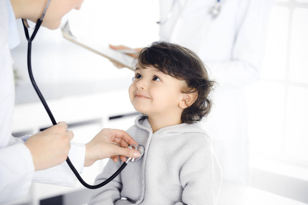Woman-doctor examining a child patient by stethoscope. Cute arab toddler at physician appointment. Medicine concept - Foto, Bild