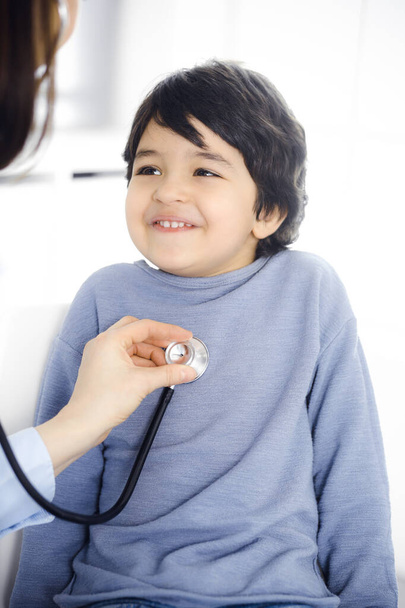 Doctor-woman examining a child patient by stethoscope. Cute arab boy at physician appointment. Medicine concept - Foto, Imagen