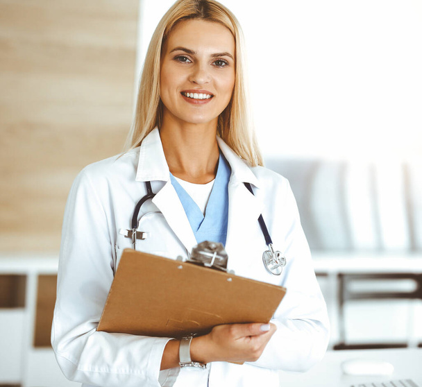 Doctor woman controls medication history records and exam results while using clipboard in sunny hospital . Medicine and healthcare concept - Photo, Image