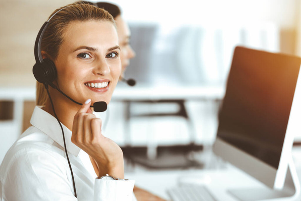 Blond female customer service representative is consulting clients online using headset in sunny office. Call center concept - Valokuva, kuva