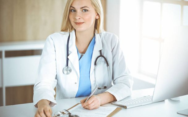Woman-doctor at work while sitting at the desk in sunny clinic. Blonde cheerful physician filling up medication history record form - Foto, Imagem
