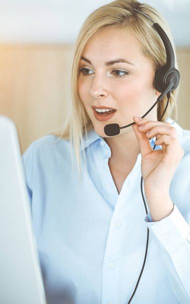 Mujer rubia de negocios sentada y comunicada por auriculares en call center en oficina soleada. Concepto de negocio de televentas - Foto, imagen