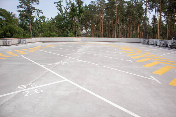 Empty rooftop parking. Directional traffic arrows and parking spots are marked with white and yellow paint on the asphalt. Parking space without cars with city skyline and park with trees - Photo, Image