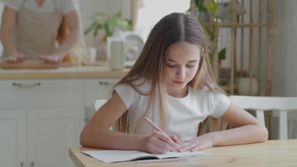 Teenager girl child schoolgirl pupil sitting at home at table in kitchen doing homework writing exercise math study learning indoor homeschooling, adult mother cooks kneads dough using rolling pin - Footage, Video