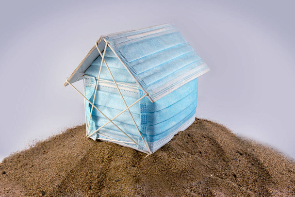 Ένα χάρτινο βιοτεχνικό σπίτι καλυμμένο με χειρουργική μάσκα στην άμμο ή την έρημο, που απεικονίζει την έννοια του ιού του κόρωνα και της πανδημικής επιδημικής απομόνωσης καραντίνας για να μείνει στο σπίτι. - Φωτογραφία, εικόνα