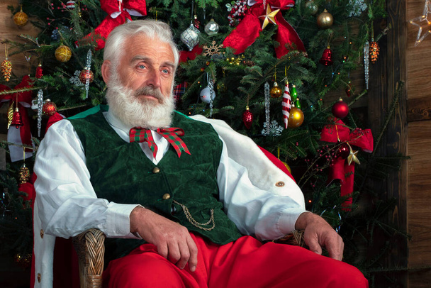 Άγιος Βασίλης στην πολυθρόνα κοντά στο χριστουγεννιάτικο δέντρο. Εορταστική εσωτερικό ξύλινο σπίτι, χαρούμενη διάθεση Πρωτοχρονιάς Πνεύμα των Χριστουγέννων. Ανώτερος άνθρωπος με πραγματική λευκή γενειάδα cosplay Άγιος Βασίλης. - Φωτογραφία, εικόνα