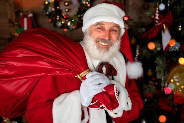 Санта-Клаус с мешком с подарками возле камина и елки. Интерьер внутри деревянного дома, новогоднее бодрое настроение. Старший мужчина с настоящей белой бородой косплей Дед Мороз. - Фото, изображение