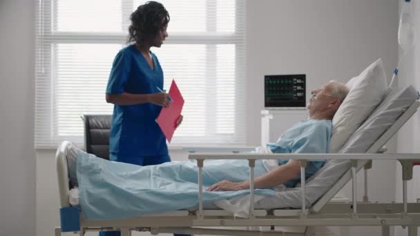 Tummaihoinen naislääkäri puhuu sairaalavuoteella makaavan vanhan miespotilaan kanssa kuntoutuksesta toipumisen jälkeen.. - Materiaali, video
