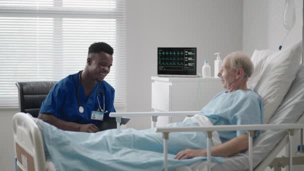 Un médico cardiólogo negro está hablando con un paciente de 60-70 años de edad, acostado en una cama en un hospital. Un neurólogo está hablando con un paciente. El paciente está conectado a una máscara de oxígeno y un dispositivo de ECG - Metraje, vídeo