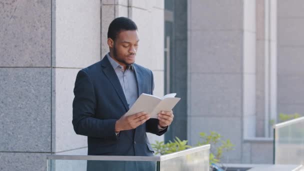 Serious confident black man wearing suit holding day planner, reading paper book or business notebook relaxing on leisure. African businessman standing on balcony of office enjoying rest during break - Footage, Video