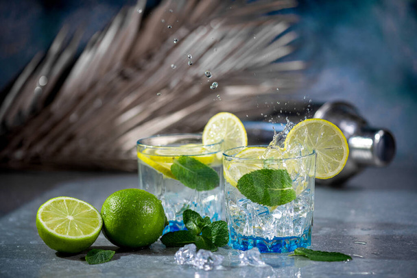 Letni koktajl alkoholowy mojito lub gin tonik z rumem, mięta, wapno i lód, narzędzia bar na betonowym szarym tle niebieski. Koktajl Mojito z pluskiem i kroplą - Zdjęcie, obraz