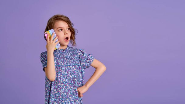 Surprized little girl holding telephone with open mouth isolated over violet color background - Photo, image