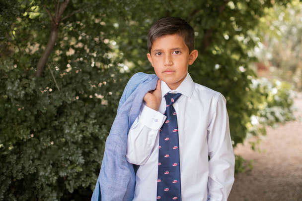 Vakava koulupoika virallisessa asussa Luotettava esiteini Latinalaisamerikkalainen poika klassisessa tyylikkäässä valkoisessa paidassa ja solmiossa, jossa on takki kädessä katsoen kameraa seisoessaan vihreässä puistossa - Valokuva, kuva