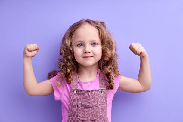 Happy caucasian little child girl showing arm muscles looking at camera isolated on purple - Foto, Imagen