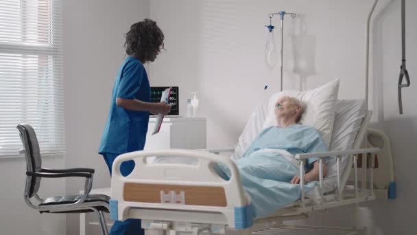 Una cardióloga negra está hablando con un paciente anciano acostado en una cama de hospital sobre rehabilitación después de la recuperación. un neurólogo de enfermedades infecciosas discute una estrategia de tratamiento - Imágenes, Vídeo