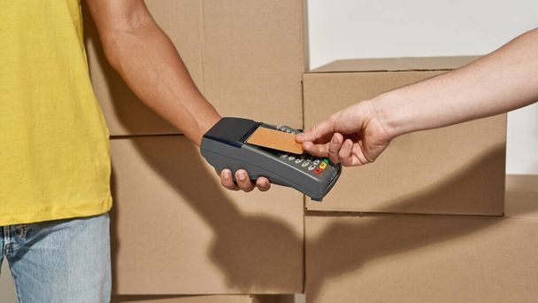 Close up shot of hands of delivery man holding terminal for a customer to pay for parcels received from courier with credit card. Cardboard boxes in the background - 写真・画像