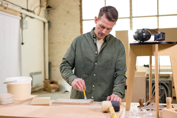 Carpenter paints wooden slat for production of furniture on table in workshop - 写真・画像