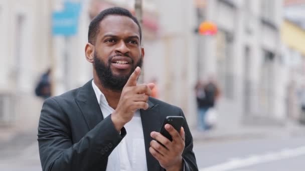 Sonriente joven biracial hombre negro sosteniendo teléfono inteligente mirando la pantalla del teléfono buscar dirección con mapa electrónico en la ciudad. Africano candidato profesional que busca empleo encuentra edificio de oficinas - Metraje, vídeo