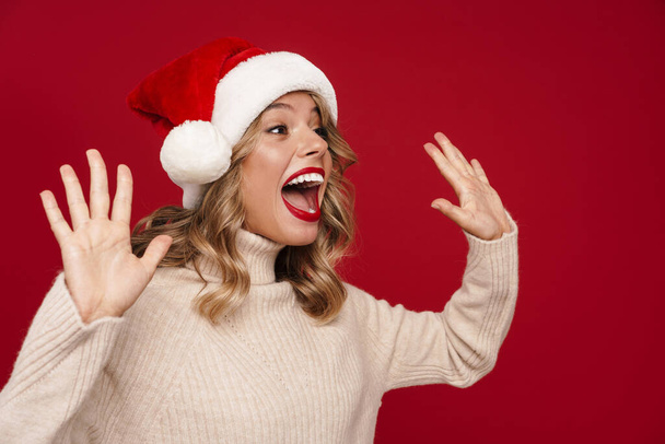 Portrait of a pretty lovely girl wearing red santa hat looking away at copy space isolated over red background, waving hand - Φωτογραφία, εικόνα
