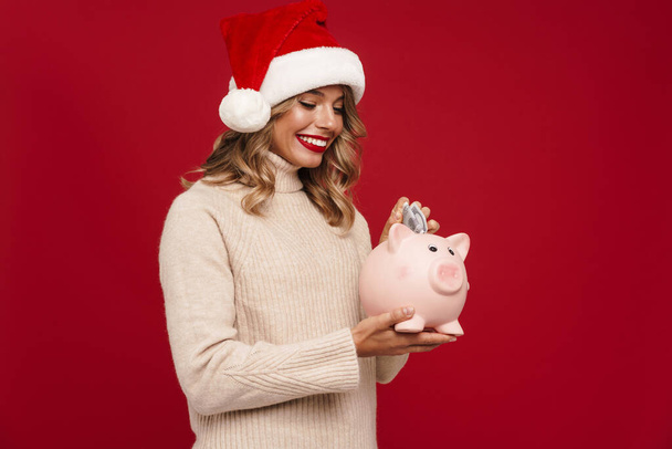 Excited pretty young woman wearing santa hat holding piggybank isolated over red background - Foto, immagini