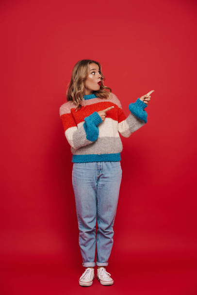 Happy young woman wearing sweater pointing away at copy space isolated over red background, full length - Фото, зображення