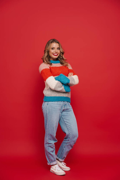 Smiling young girl wearing sweater standing isolated over red background, full length - Фото, зображення