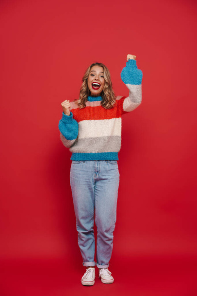 Smiling young girl wearing sweater standing isolated over red background, full length, celebrating success - Foto, immagini