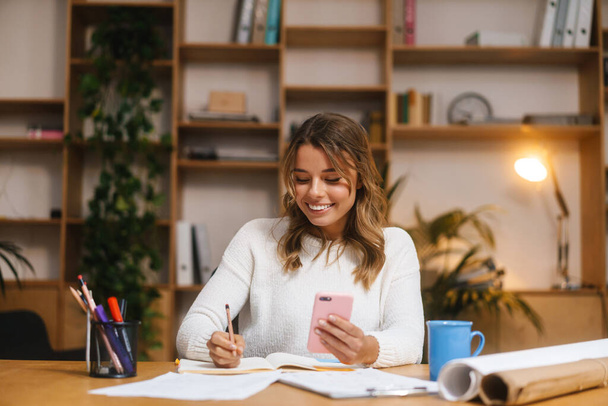 Smiling attractive young woman entrepreneur using mobile phone while working with documents at the office desk - Foto, imagen