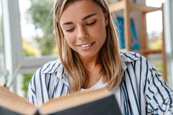 Gülümseyen çekici genç bayan tasarımcı atölyede otururken kitap okuyor. - Fotoğraf, Görsel