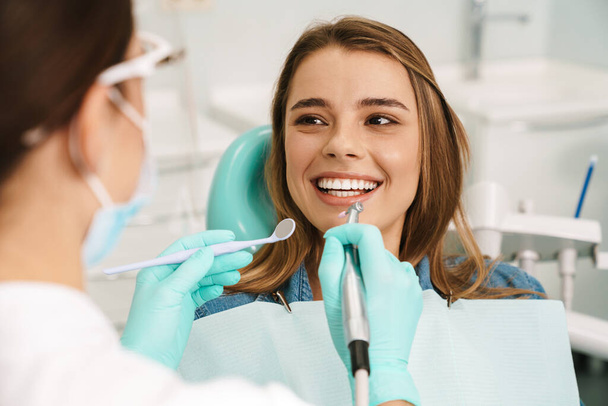 European smiling woman sitting in medical chair while dentist fixing her teeth at dental clinic - 写真・画像