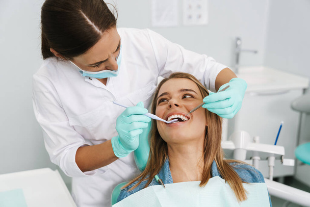 European young woman sitting in medical chair while dentist fixing her teeth at dental clinic - Zdjęcie, obraz