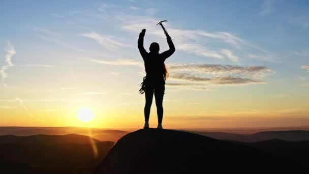 Langzame beweging van een vrouwelijke klimmer overwinnend steekt haar handen omhoog met een ijsbijl terwijl ze op de top van een berg staat bij zonsondergang - Video