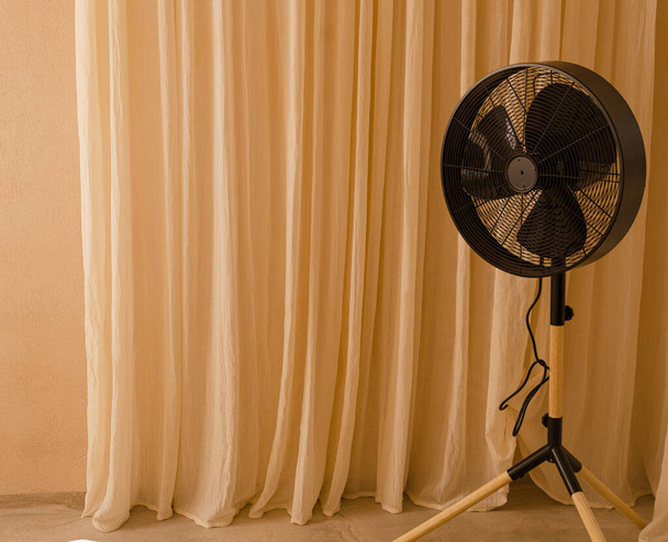 A big black fan standing in front of beige curtains. Minimal summer heat composition. Retro air condition concept - Photo, Image