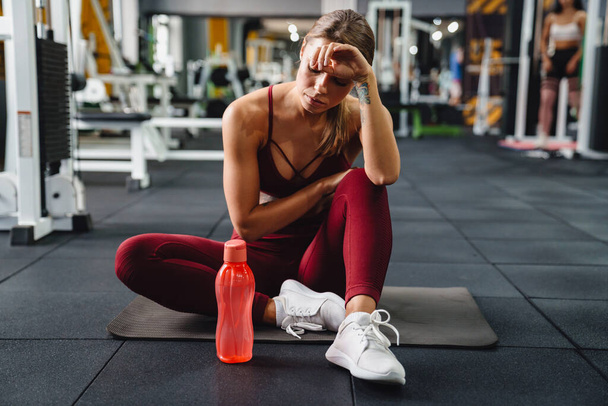 Tired athletic sportswoman sitting on mat while working out in gym - Photo, image