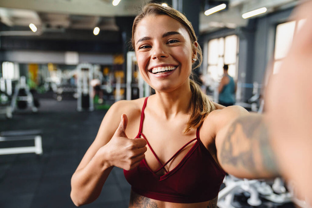 Smiling athletic sportswoman showing thumb up while taking selfie photo in gym - Foto, Imagem