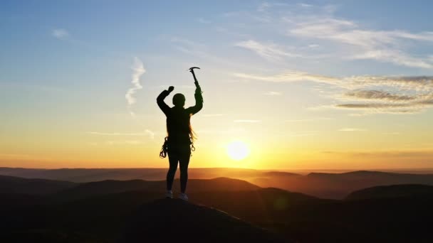 Een jonge vrouwelijke klimmer heft haar handen overwinnend op terwijl ze bij zonsondergang op de top van de berg staat. - Video