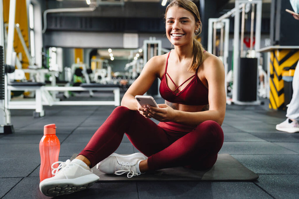 Photo of cheery positive young woman fitness coach using mobile phone in gym - Fotografie, Obrázek