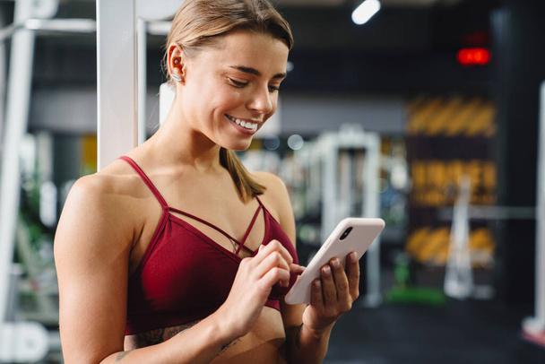 Photo of cheery positive young woman fitness coach using mobile phone in gym - Fotografie, Obrázek