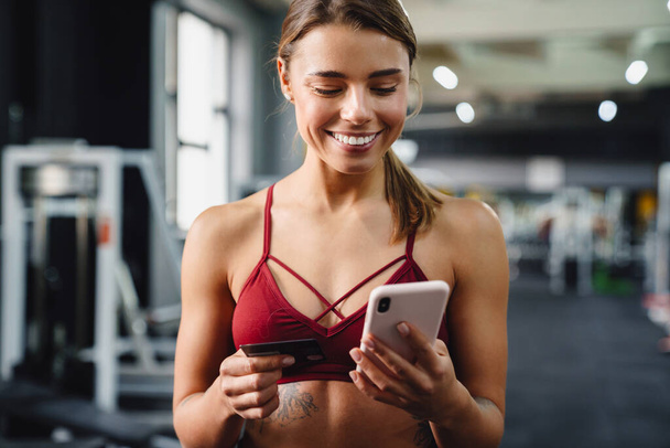 Foto de una joven sonriente entrenadora de fitness usando un teléfono móvil en el gimnasio mientras sostiene la tarjeta de crédito - Foto, imagen