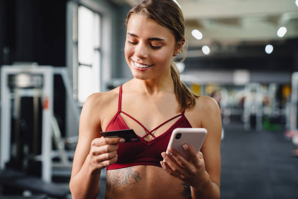 Photo of smiling young woman fitness coach using mobile phone in gym while holding credit card - Valokuva, kuva
