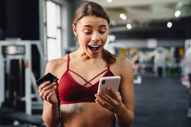 Photo of shocked excited young woman fitness coach using mobile phone in gym while holding credit card - Zdjęcie, obraz