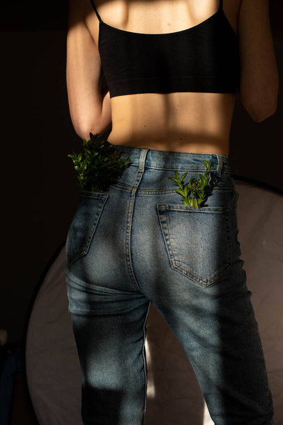 ジーンズのポケットに緑の植物。ストライプの形で体に影のパターンを持つ背中の美しい若い女の子の美しさの肖像画。ファッション美。拒食症の女の子が背を向けて. - 写真・画像