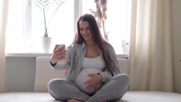 Tabletli mutlu hamile bir kadın, cep telefonuyla arkadaşlarıyla sohbet ederek kocasıyla akıllı telefonlardan iletişim kuruyor. gebelik, teknoloji, tıp - Video, Çekim