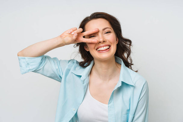 Porträt einer lächelnden Frau, die ein Siegeszeichen zeigt und isoliert in eine Kamera auf weißem Hintergrund blickt. - Foto, Bild